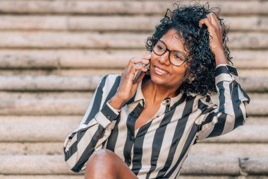 Latin Afrikalı Amerikalı kadın sokakta cep telefonuyla oturuyor.