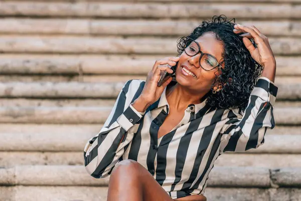 Latin Afrikalı Amerikalı kadın sokakta cep telefonuyla oturuyor.