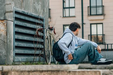 Sırt çantası ve bisikletiyle sokakta oturan cep telefonu olan genç bir adam ya da öğrenci.