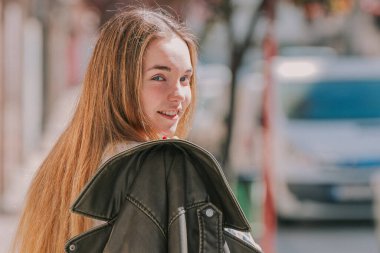 Genç, sarışın, uzun saçlı, sokakta sırtında ceketle dolaşan bir kız.