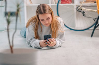 Sarışın kız evde oturmuş cep telefonuna bakıyor.