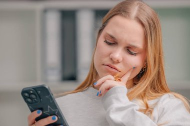 Evde cep telefonu olan sarışın bir kız ve şüpheli ve düşünceli bir yüz ifadesi var.