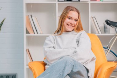 Gülümseyen sarışın genç kadın evdeki koltukta oturmuş kameraya mutlu bir şekilde bakıyor.