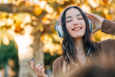 Sonbaharda açık havada şapkalı gülümseyen genç bir kadın kulaklık ve telefonla müzik dinliyor.