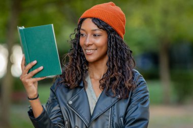 Sonbaharda şapkalı genç bir kadın elinde kitapla gülümsüyor.