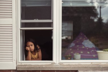 Çocuk pencereden dışarı bakıyor, yalnız sıkılmış ve üzgün.