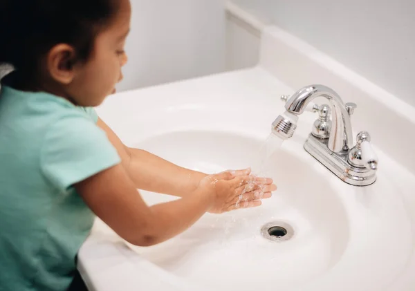 Diverse pre school girl at home in bathroom cleaning sink and washing hands, chores and hygiene