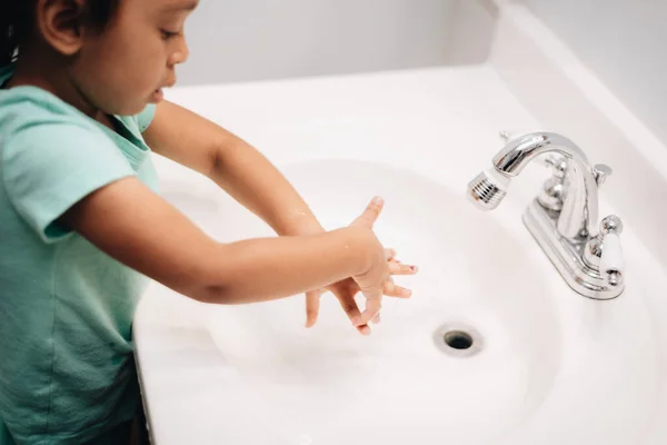 Diverse pre school girl at home in bathroom cleaning sink and washing hands, chores and hygiene