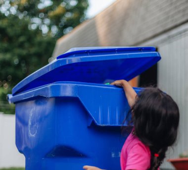 Diverse pre school girl putting glass jar into recycling bin, environmentally friendly, consciou