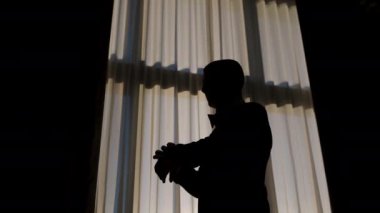 Silhouette of a man putting on his jacket in dark room on the background of the window. High quality 4k footage