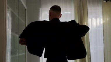 Silhouette of a man putting on his jacket in dark room on the background of the window. High quality 4k footage