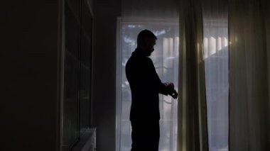Silhouette of a man putting on his jacket in dark room on the background of the window. High quality 4k footage