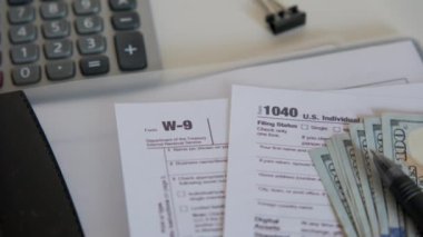 An American dollar tax bill, pen calculator and U.S. money. 1040 and W9 individual tax form are placed on desk of American citizen. High quality 4k footage