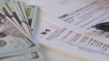 An American dollar tax bill, pen calculator and U.S. money. 1040 and W9 individual tax form are placed on desk of American citizen. High quality 4k footage