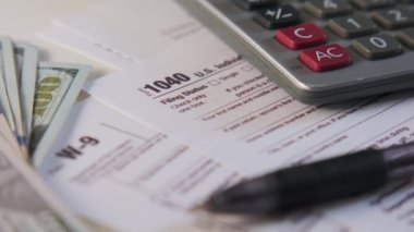 An American dollar tax bill, pen calculator and U.S. money. 1040 and W9 individual tax form are placed on desk of American citizen. High quality 4k footage