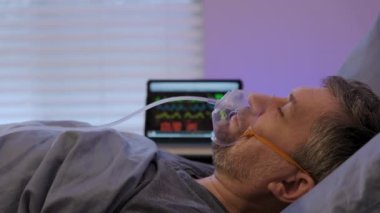 Side view of mature Caucasian man wearing oxygen mask, lying in bed in room of clinic,. High quality 4k footage