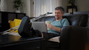 A happy man with a broken leg is working on a laptop. High quality 4k footage