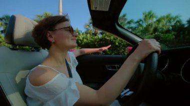 Mutlu kadın gün batımında el sallayarak Cabriolet araba kullanıyor. Yan görüş. Yüksek kalite 4k görüntü