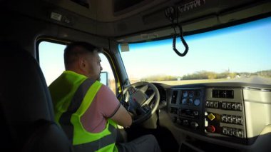 Güvenli yelek giyen genç yakışıklı yarı profesyonel kamyon şoförü güneşli bir günde bir kamyona biniyor. Kamyonun içinden vurulmuş. Yüksek kalite 4k görüntü