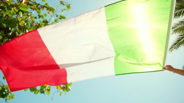 Açık mavi gökyüzü ve tropikal ağaçlardan oluşan güçlü bir rüzgara karşı İtalyan bayrağını tutan kişi. Yüksek kalite 4k görüntü