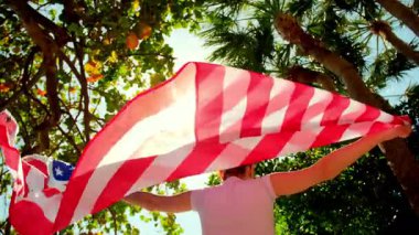 Arkaplanı açık mavi gökyüzü, deniz ve tropikal ağaçlardan oluşan güçlü bir rüzgara karşı ABD bayrağı tutan bir kadın. Yüksek kalite 4k görüntü