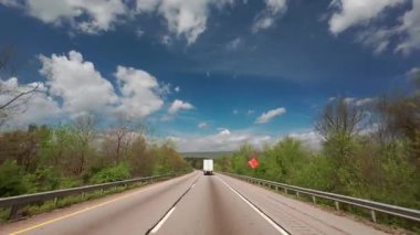 POV boş bir otoyolda ilerliyor. Önünde parlak mavi bir gökyüzünün altında beyaz bir kamyon var. Her iki tarafında da kabarık bulutlar ve yeşil bahar ağaçları var. Yüksek kalite 4k görüntü