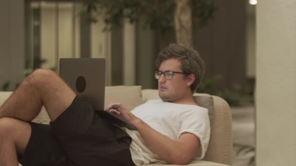 全神贯注的男人穿着白色T恤 用笔记本电脑发短信 靠在家里舒适的沙发上 从事计算机 远程工作和连接等方面工作的商人 自由职业者的概念 — 图库视频影像
