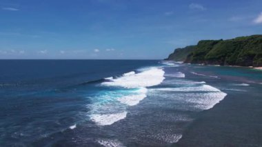 Yavaş çekim drone çekimi geriye doğru uçan okyanus dalgaları, yeşil tepeler ve deniz kıyısında mavi gökyüzü. Güzel doğanın sinematik üst görüntüsü. Tatil kavramı