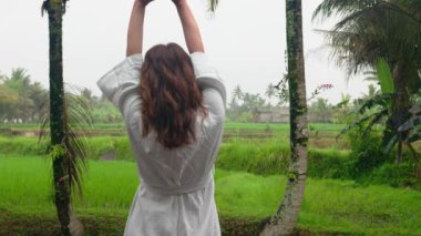 Gecelikli bir kadının uyanıp ellerini kaldırarak Bali, Endonezya 'daki tropikal palmiyelere ve yeşil pirinç tarlalarına baktığı sahne. Seyahat, tatil ve otel kavramı