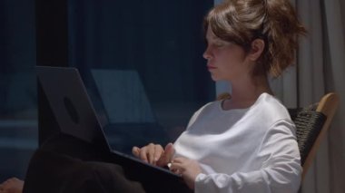Koltuğunda oturan düşünceli genç bir kadın, dizüstü bilgisayarda kayan bir yan görüş. Akşamları evden çalışan düşünceli bir iş kadını. İnternet ve serbest meslek anlayışı