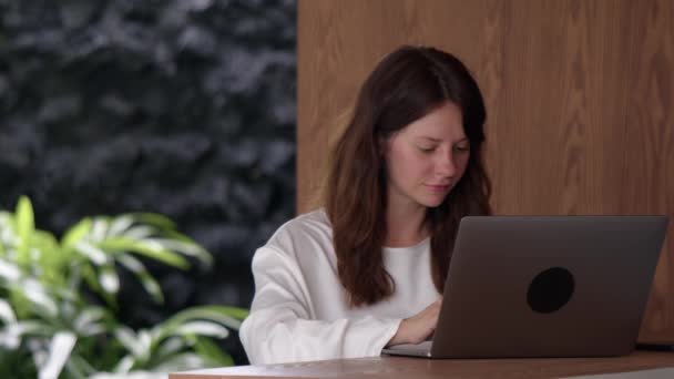 穿着白色衬衫的微笑女人用笔记本电脑发短信 侧面看木制台面 在网吧或酒吧里进行在线交流 远程工作和自由职业的概念 — 图库视频影像