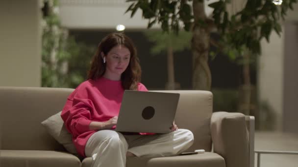 穿着粉色运动衫 穿着笔记本电脑打字的漂亮女人微笑着 自由职业者坐在沙发上 用电脑和耳机 在线通信和远程工作的概念 — 图库视频影像