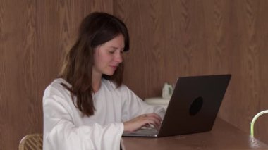 Beyaz tişörtlü çekici genç bir kadın laptopta mesaj atıyor, yan manzara ahşap duvar ve masa. İnternette sörf yapmak ve gezinmek. Çevrimiçi iletişim, aygıt ve bilgisayar kavramı