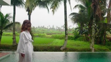 Endonezya, Bali 'de havuzun kenarında yürüyen gecelikli rahat genç bir kadın. Yükselen tarlalar, palmiye ağaçları, güzel doğa ve lüks otel. Seyahat ve tatil kavramı