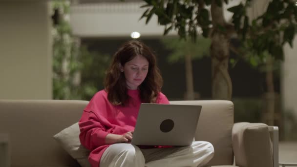 漂亮的年轻女子坐在沙发上 在酒店大堂的笔记本电脑上打字模糊了背景 浏览互联网和网上购物 数字网络和通信的概念 — 图库视频影像