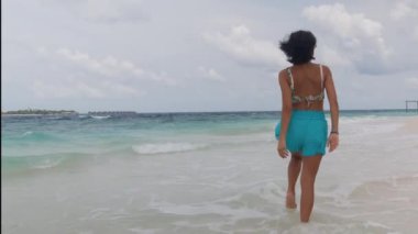 Arkadan uzun bir görüntü, mavi şortlu bir kadın Maldivler 'de okyanus kumsalında yürüyor. Yaz tatilinin tadını çıkaran bir kadın dalgalara basıyor. Seyahat, turizm, seyahat konsepti. Yüksek