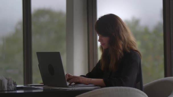 沉着而美丽的黑发女人 穿着黑色衬衫 在笔记本电脑上打字 侧观树木的全景窗 在线交流 社交媒体和互联网的概念 — 图库视频影像