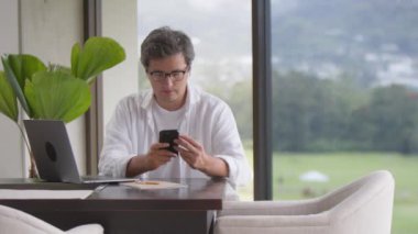 Beyaz gömlekli gülümseyen bir işadamı telefonda yazıyor, rahat bir evde oturuyor, güzel manzaranın panoramik penceresinde oturuyor. Mesafe ve online alışveriş kavramı
