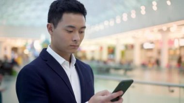 Asyalı genç iş adamı portresi. Takım elbise giymiş, kahve içen, elinde telefon tutan ve iş merkezinde yürüyen siyah saçlı.
