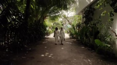 Beyaz gelinlik giymiş iki güzel kadın köyde ve Maldivler 'deki tropikal orman adasında sahile doğru yürüyorlar. Tatil ve seyahat kavramı