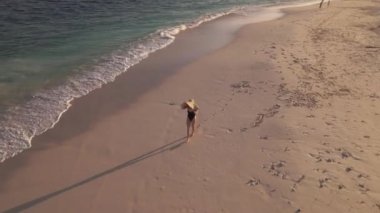 Maldivler 'de güneş şapkası, kumlu plaj, siluet ve turkuaz Hint okyanusu dalgaları içinde yürüyen genç bir kadının hava görüntüsü. Turizm ve hafta sonu kavramı.