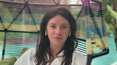 Beyaz bornozlu çekici bir kadının portresi müzik dinliyor terasta kablosuz kulaklık kullanıyor, arka planda orman var. Havuz salıncağında dinleniyorum. Mesafe çalışması kavramı