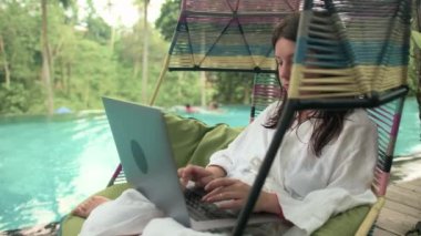 Beyaz bornozlu çekici bir kadın sonsuzluk havuz terasında dizüstü bilgisayar kullanıyor arka planda orman, bilardo salıncağında serinliyor. Mesafe çalışması kavramı