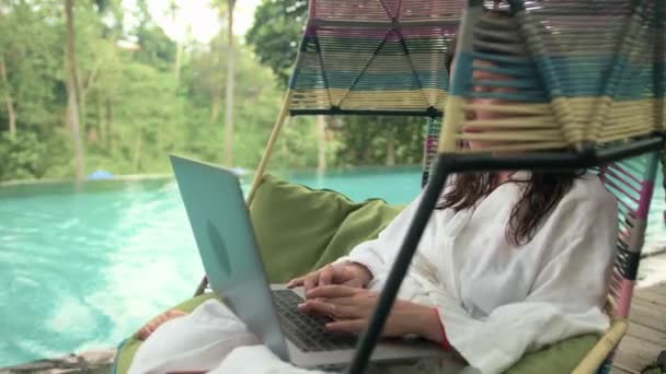 一个穿着白色浴衣的漂亮女人在无限大的游泳池平台上使用笔记本电脑 背景是丛林 泳池荡秋千让人胆寒 远程工作的概念 — 图库视频影像