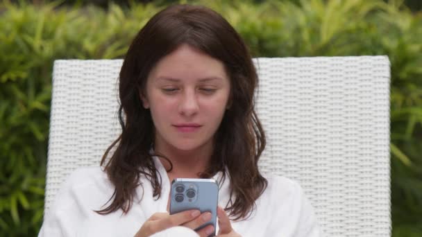 テラスでスマートフォンを使用した白いバスローブの魅力的な女性の肖像画 背景にジャングル 距離作業という概念 ソーシャルメディアのコミュニケーション — ストック動画