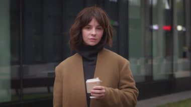 Kahverengi ceketli kadın kahve fincanı tutuyor, sokak tarzı, cam duvarın yanında, rüzgarlı bir günde modern yaşam tarzı konsepti.