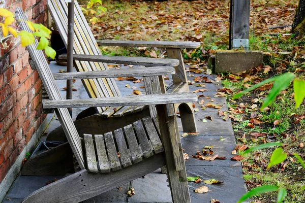 An image of two old wooden patio chairs on a small stone patio surrounded by colorful autumn leaves.