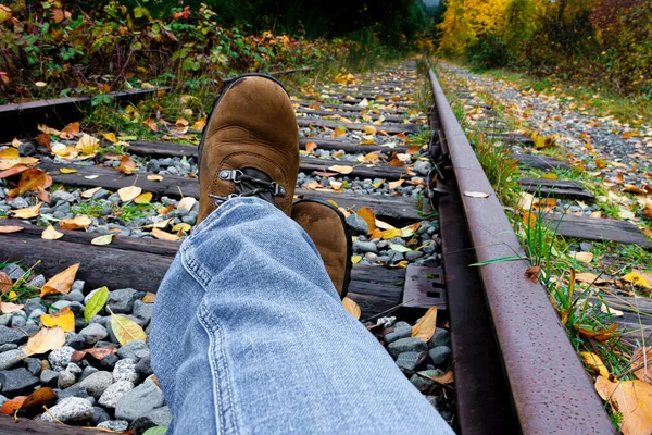 An image of a pair of stretched legs with tan hiking boots resting on old abandoned railway tracks covered with bright autumn leaves.