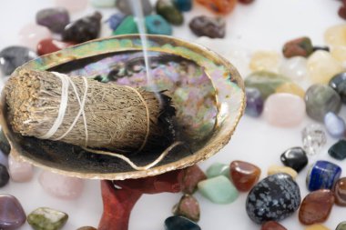 Beyaz bir masanın üzerinde küçük iyileştirici kristallerle yanan bir adaçayı lekesi resmi.. 