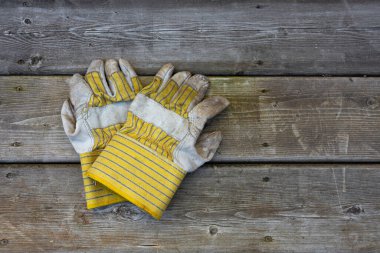 Eski sarı ve gri iş eldivenleri, aşınma ve kir izleri gösteriyor, eski ve yıpranmış ahşap bir yüzeyde yatıyor. Eldivenlerde bilek bölgesinde çizgili bir desen var.. 